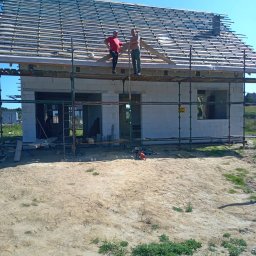 Usługi ciesielskie i wykończeniowe Wojciech Lesner - Dwóch mężczyzn na rusztowaniu montuje drewnianą konstrukcję dachu na surowym budynku z białych bloczków, piła łańcuchowa leży na ziemi.