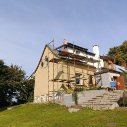 Usługi ciesielskie i wykończeniowe Wojciech Lesner - Prace na wysokościach: Dwóch mężczyzn na rusztowaniu przy żółtym domu, przygotowujących się do prac na elewacji, widoczne ślady wcześniejszych napraw i odpadający tynk.