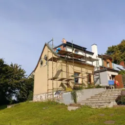 Prace na wysokościach: Dwóch mężczyzn na rusztowaniu przy żółtym domu, przygotowujących się do prac na elewacji, widoczne ślady wcześniejszych napraw i odpadający tynk.