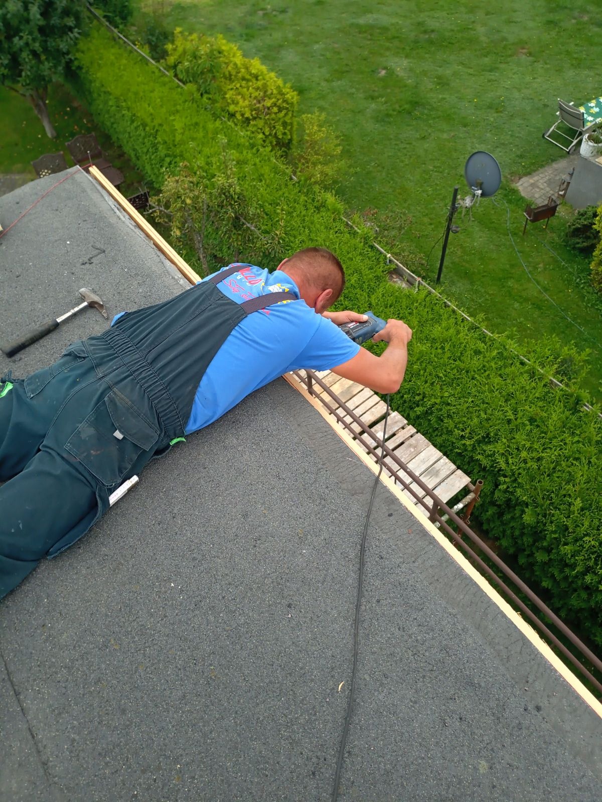 Pracownik w niebieskiej koszulce i granatowym kombinezonie mocuje drewniane elementy konstrukcji na dachu pokrytym papą, widok z góry na zielony ogród z krzewami i satelitą.