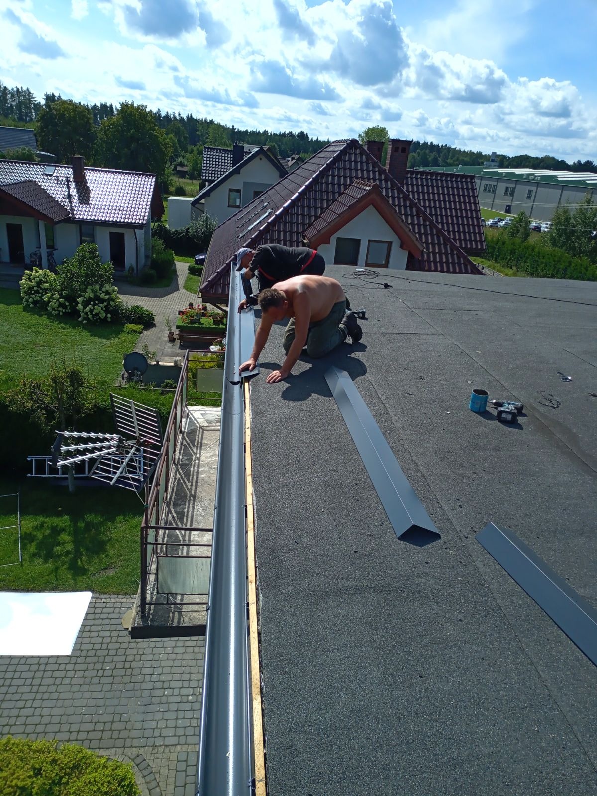 Montaż metalowej rynny dachowej na papie przez dwóch mężczyzn, jeden bez koszulki, z widocznymi narzędziami i elementami rynny na dachu domu jednorodzinnego w słoneczny dzień.