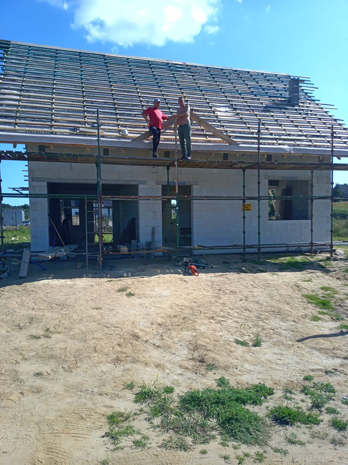 Dwóch mężczyzn na rusztowaniu montuje drewnianą konstrukcję dachu na surowym budynku z białych bloczków, piła łańcuchowa leży na ziemi.