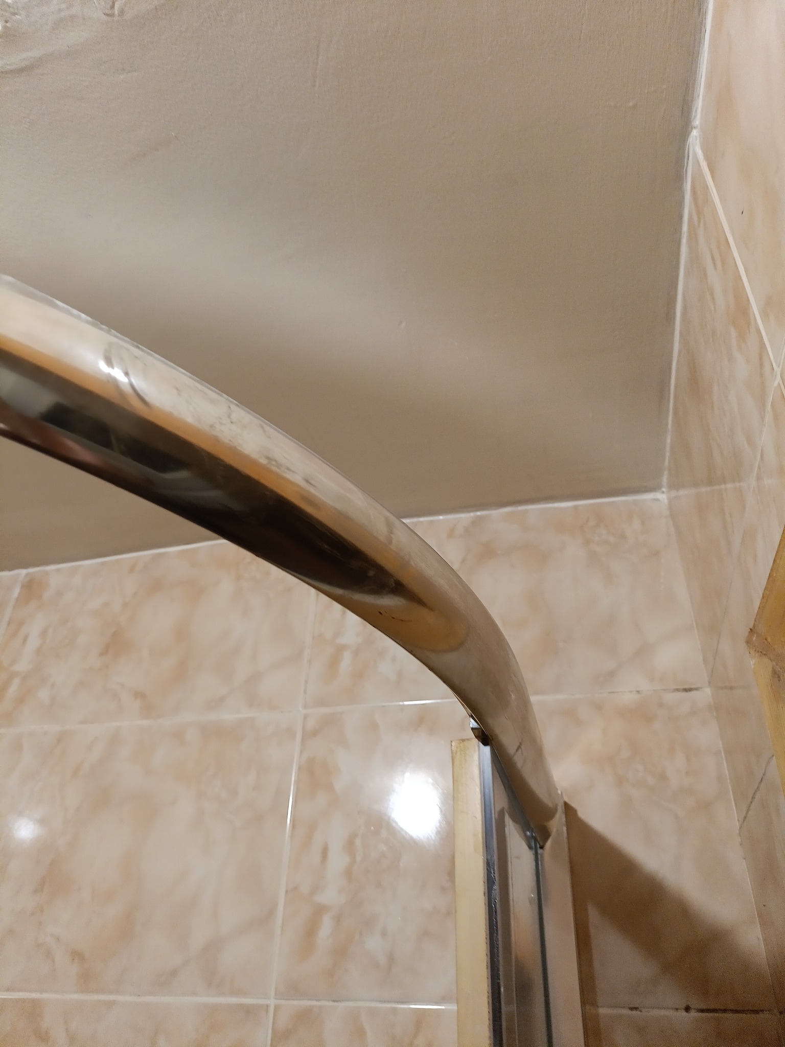 Fragment kabiny prysznicowej z chromowaną ramą na tle beżowych płytek i sufitu z widocznymi niedoskonałościami.