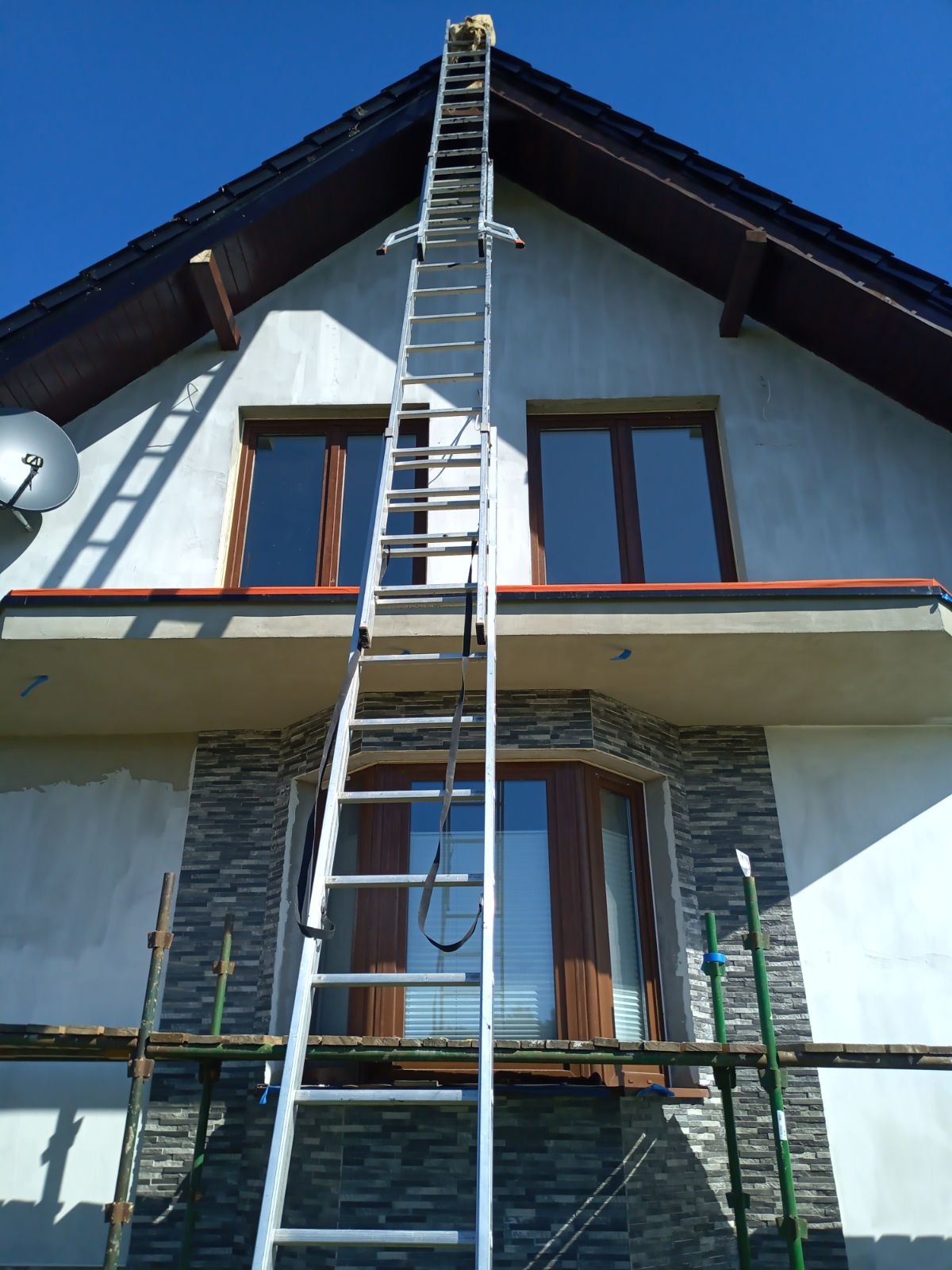 Dom w trakcie prac wykończeniowych elewacji, widoczna długa drabina oparta o dach i rusztowanie przy ścianie, ściany częściowo otynkowane, z fragmentami wykończonymi kamieniem dekoracyjnym, okna...