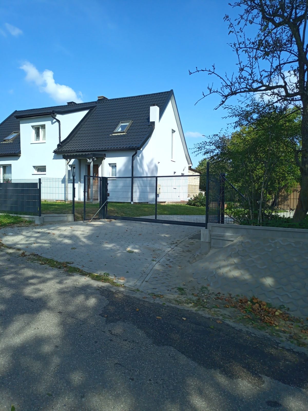 Nowoczesny, biały dom jednorodzinny z czarnym dachem i metalowym ogrodzeniem z bramą przesuwną, widok z ulicy w słoneczny dzień.