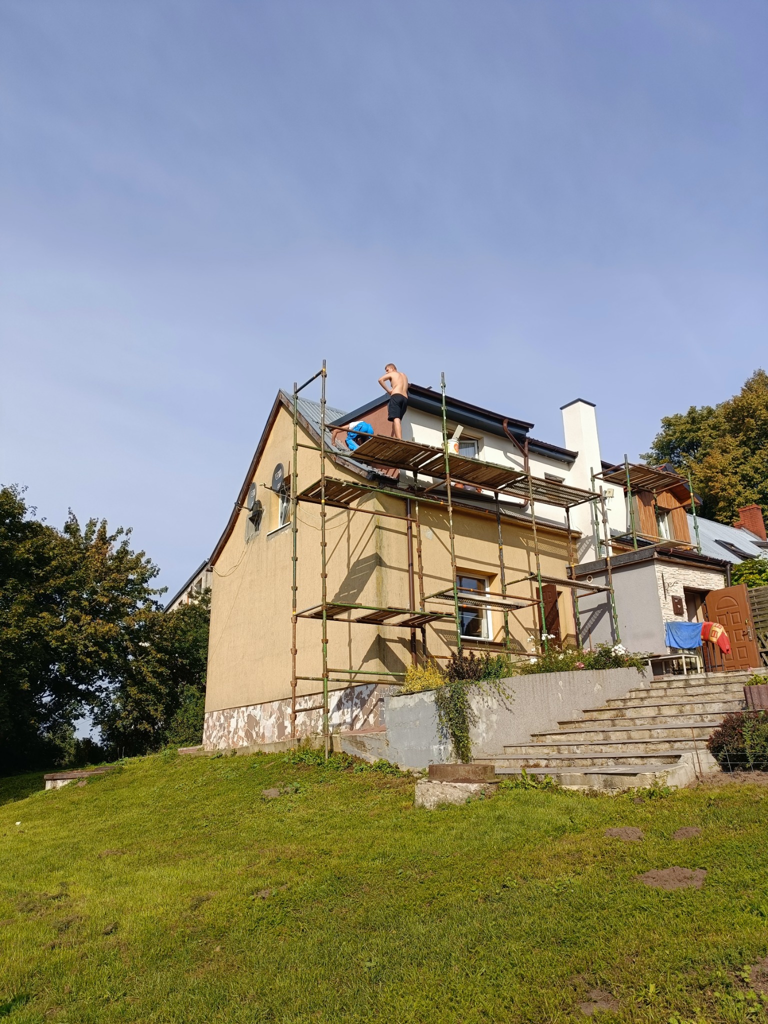 Prace na wysokościach: Dwóch mężczyzn na rusztowaniu przy żółtym domu, przygotowujących się do prac na elewacji, widoczne ślady wcześniejszych napraw i odpadający tynk.