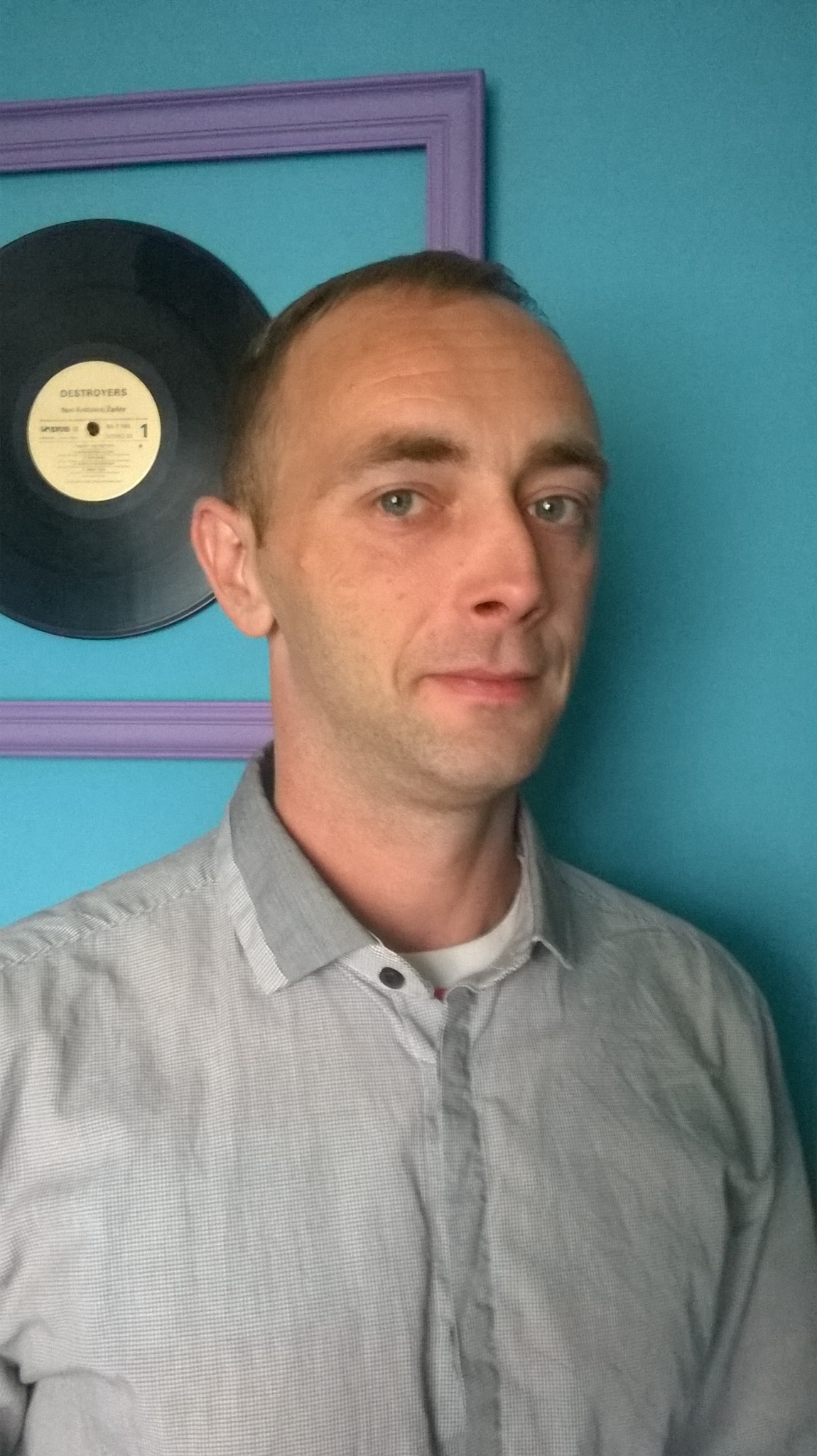 Portret mężczyzny w szarej koszuli na tle turkusowej ściany z fioletową ramką i płytą winylową.
