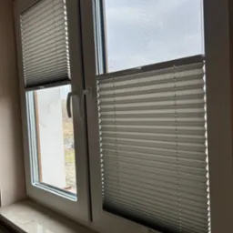 Okno z białymi ramami, na jednym skrzydle zamontowana moskitiera, na drugim szare plisy w połowie zasłonięte.