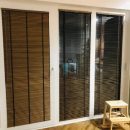 Okno balkonowe z brązowymi przesłonami okiennymi, częściowo zasłoniętymi, z drewnianą drabinką stojącą na parkiecie w jodełkę.