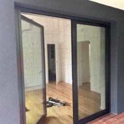 Rozsunięta moskitiera plisowana w kolorze czarnym zamontowana w drzwiach balkonowych, widoczny drewniany parkiet wewnątrz i narzędzia montażowe na podłodze.