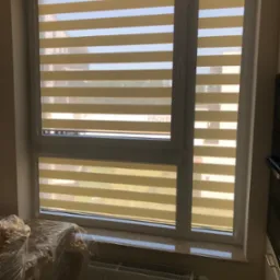 Beżowo-białe, poziome pasy na oknach w nowoczesnym domu, widoczny fragment mebla zabezpieczonego folią.