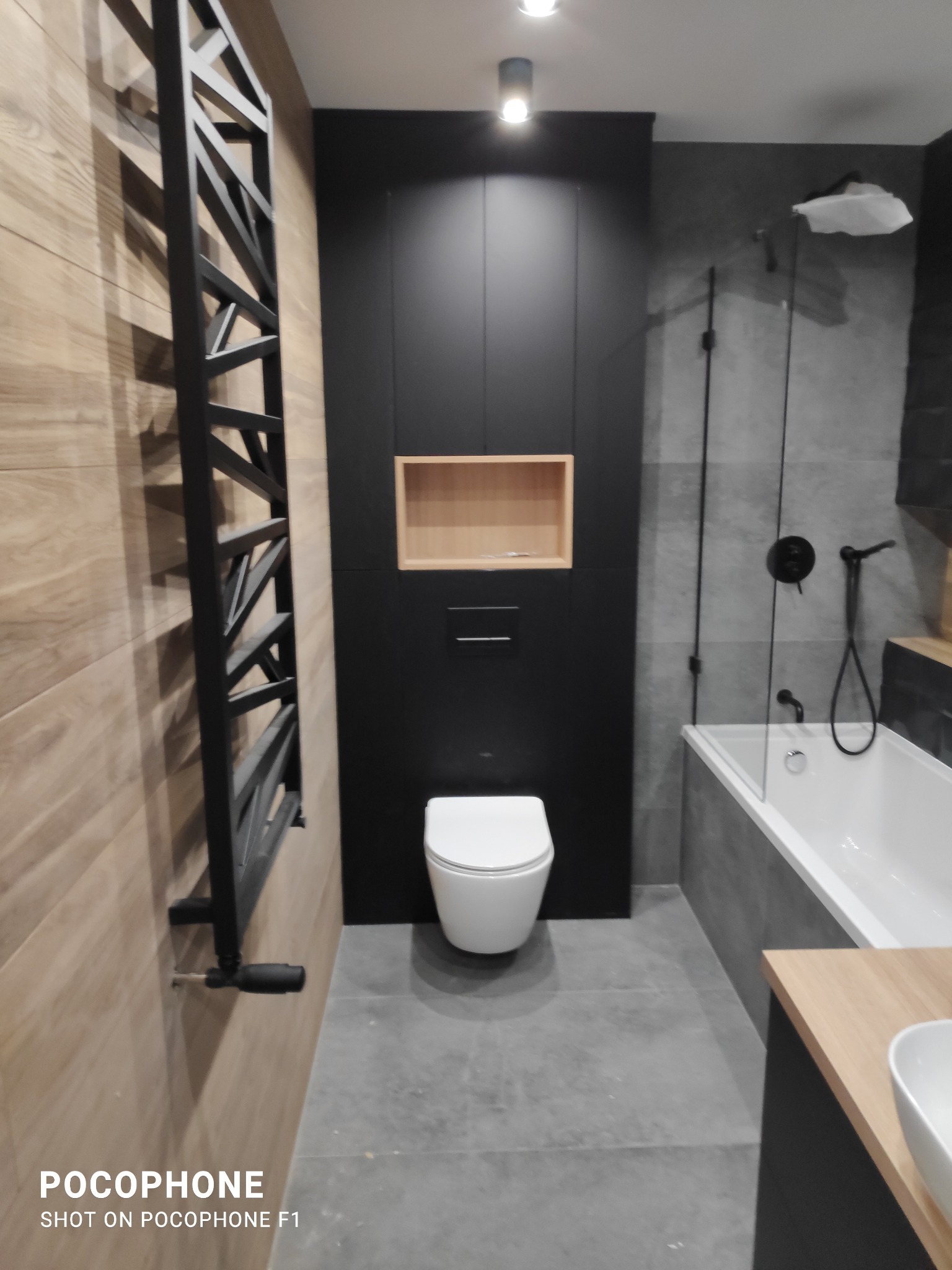 Nowoczesna łazienka z drewnianymi akcentami, czarnymi matowymi elementami i grzejnikiem drabinkowym na tle płytek imitujących drewno.