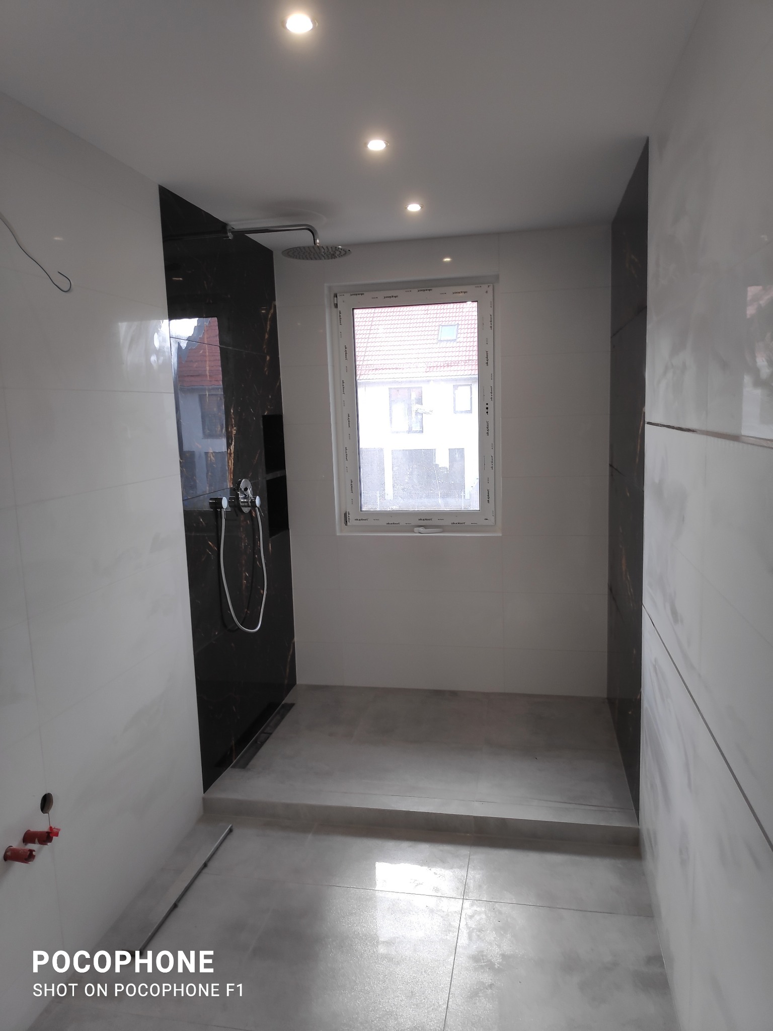 Nowoczesna łazienka z kabiną prysznicową typu walk-in, wykończona białymi i czarnymi płytkami imitującymi marmur, widoczny odpływ liniowy i okno z widokiem na budynek mieszkalny.