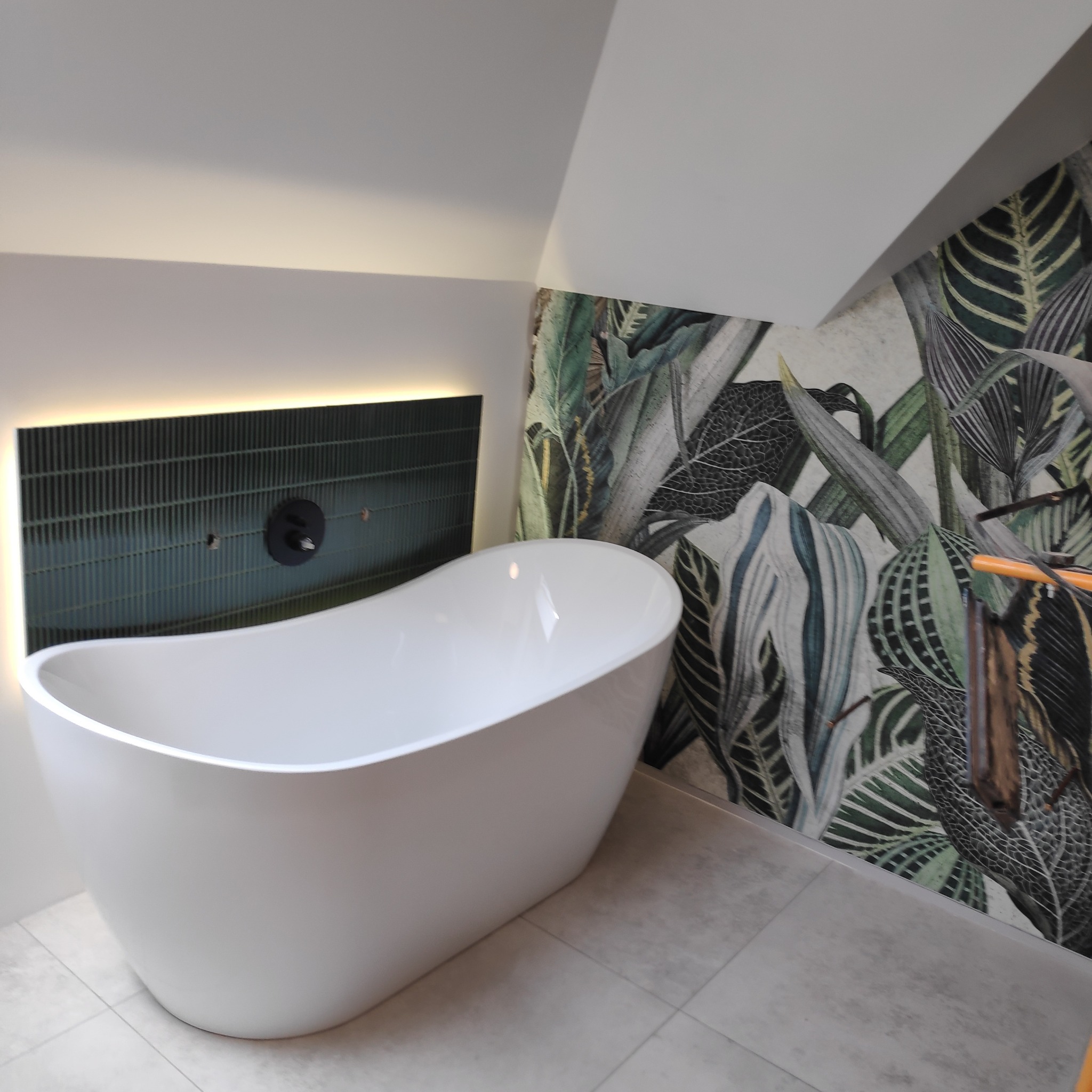 Wolnostojąca wanna owalna na tle ściany z motywem roślinnym i podświetlonej, ciemnozielonej ściany z armaturą.