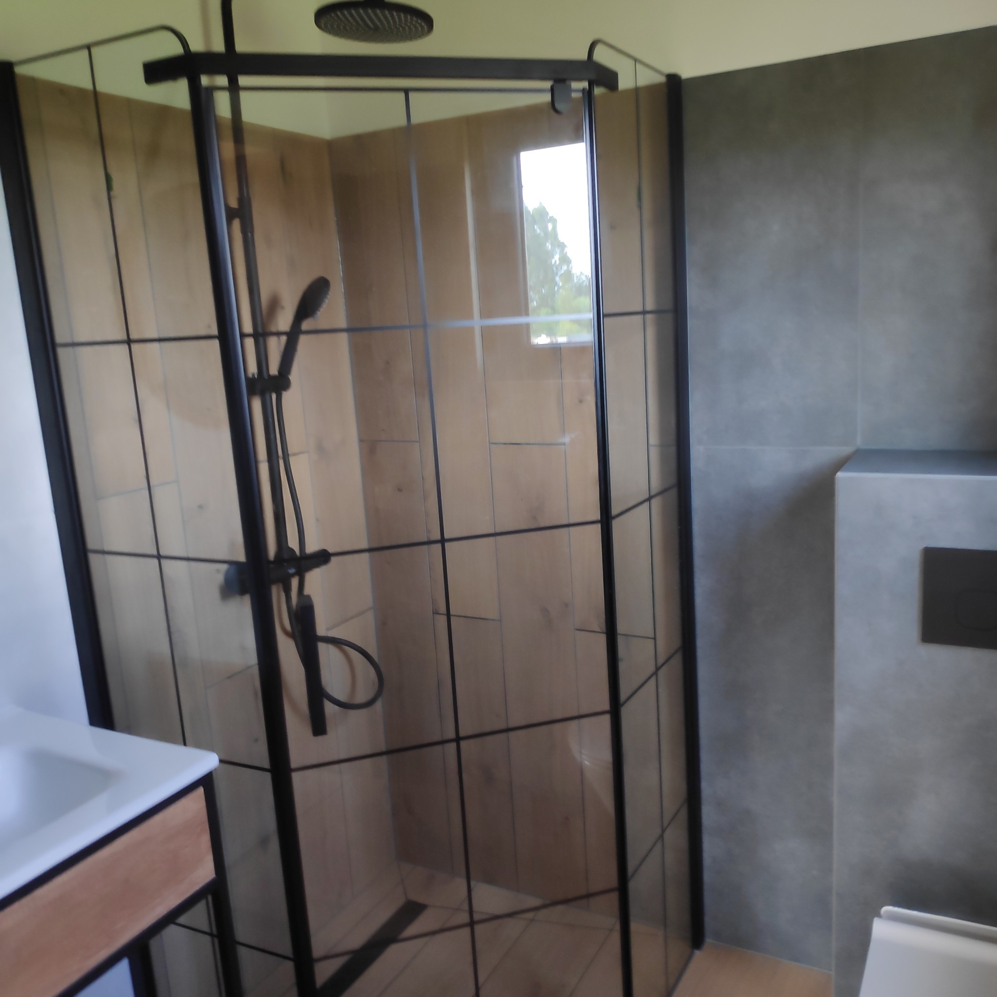 Nowoczesna kabina prysznicowa z czarnymi profilami i deszczownicą, drewnopodobne płytki na ścianach, szara ściana z ukrytym systemem spłukiwania toalety, umywalka z szafką.