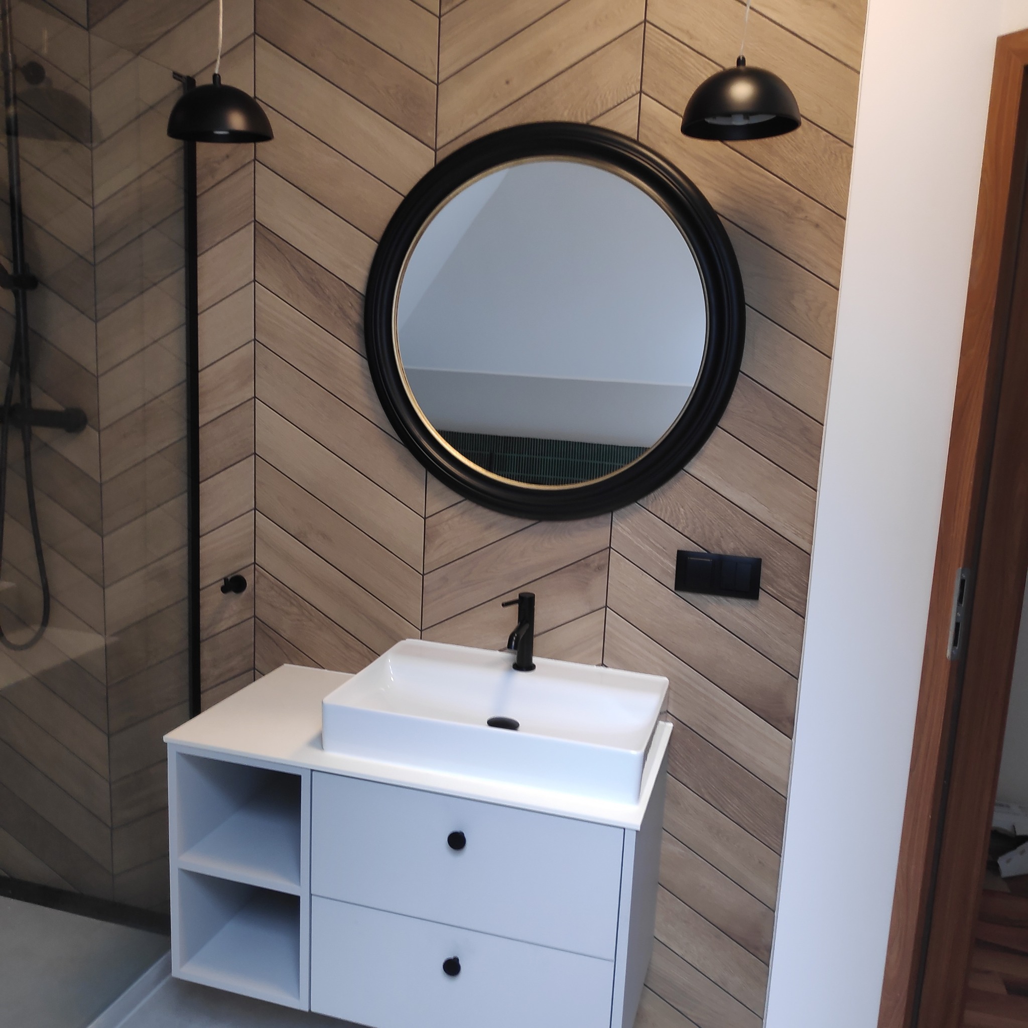 Nowoczesna łazienka z drewnopodobnymi płytkami ułożonymi w jodełkę, okrągłym lustrem w czarnej ramie, białą szafką pod umywalką z czarną armaturą i wiszącymi lampami.