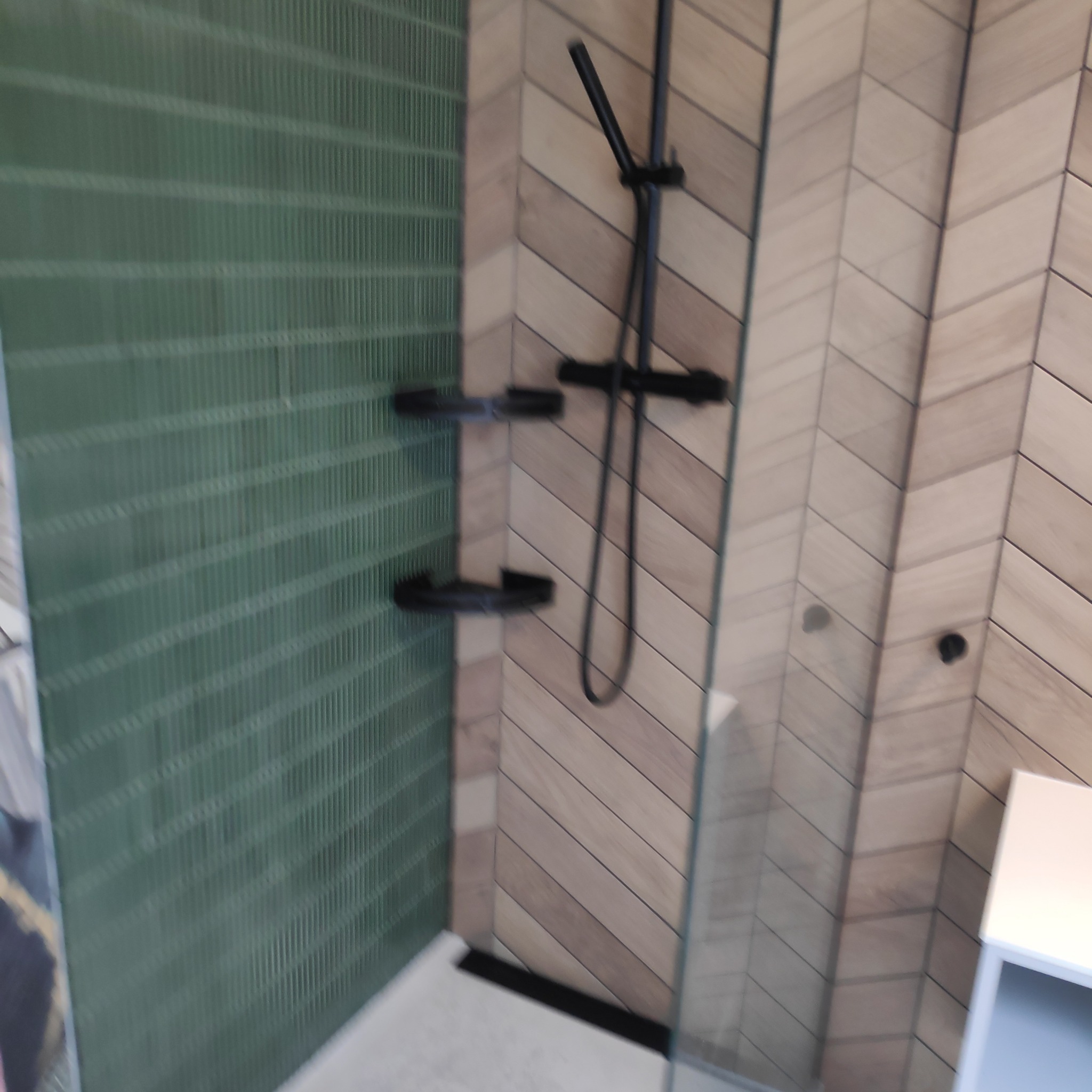 Nowoczesna kabina prysznicowa z zieloną ścianą z płytek i armaturą w kolorze czarnym, wykończona płytkami ułożonymi w jodełkę.