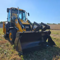 Żółta koparko-ładowarka JCB 3cx Plus z 2023 roku, zaparkowana na trawiastym terenie w Warszawie, z kontenerami w tle.