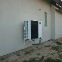 Zewnętrzna jednostka pompy ciepła zamontowana na ścianie budynku w trakcie wykańczania, widoczne niedoskonałości elewacji i nierówny teren wokół.