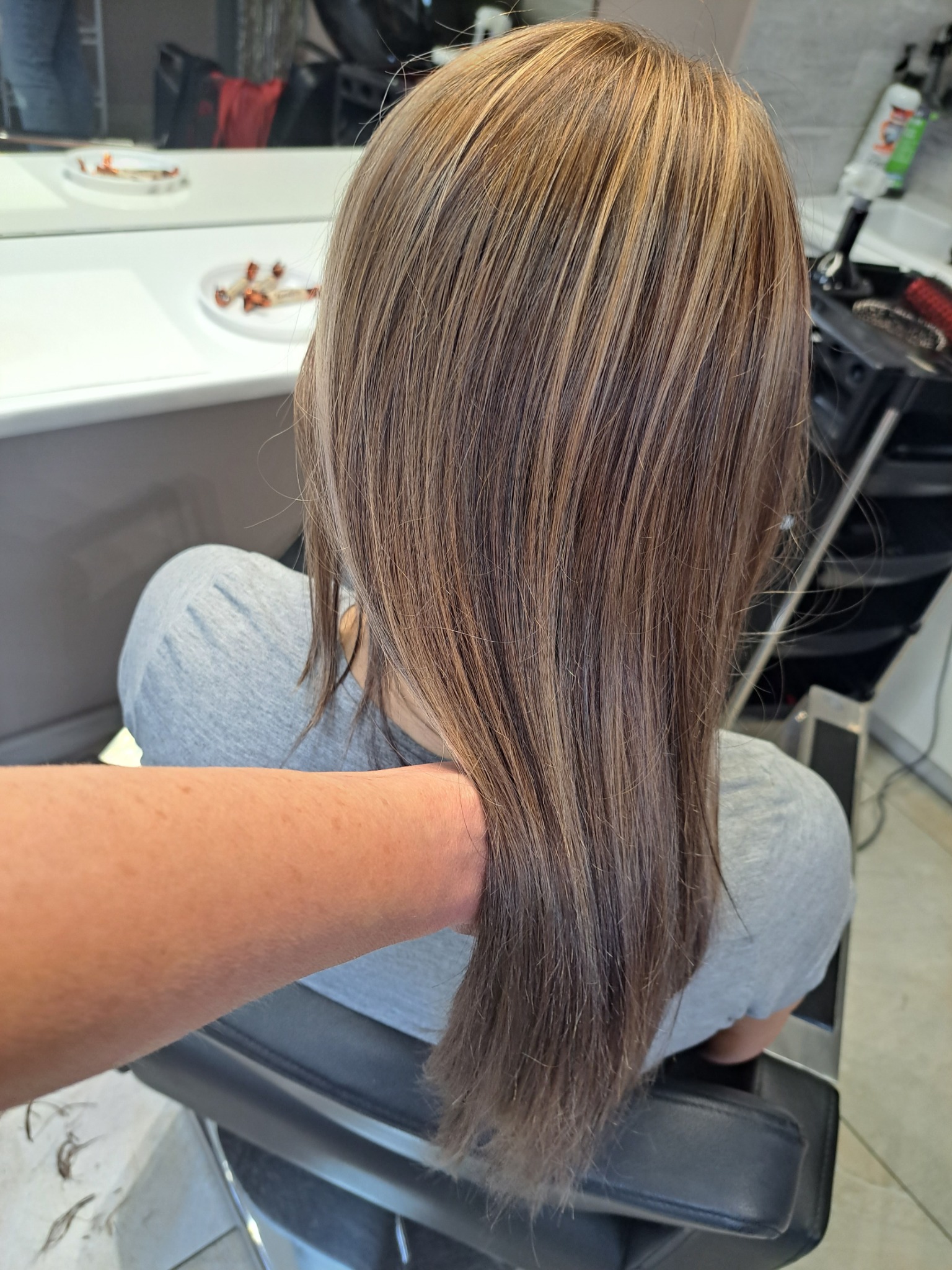 Długie, proste włosy z refleksami, po koloryzacji, widok z tyłu, w salonie, ręka stylisty przytrzymuje włosy.