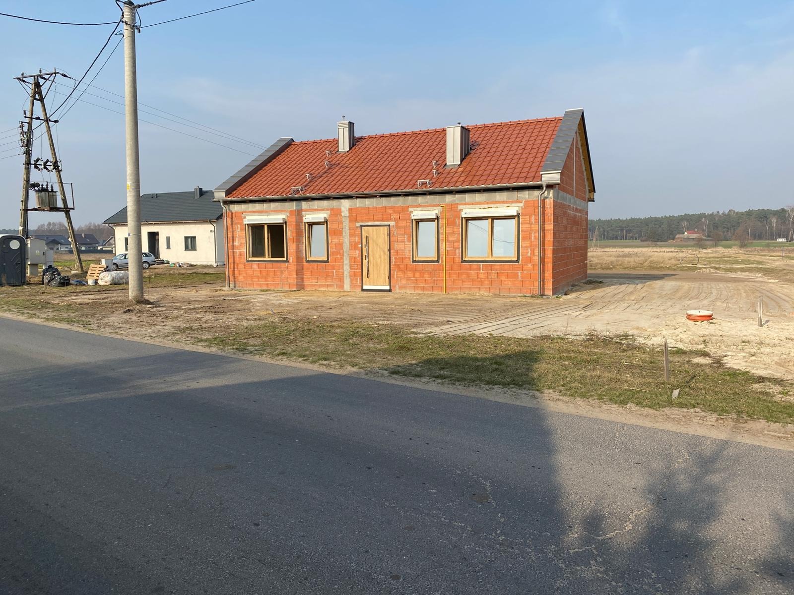 Surowy, parterowy dom z czerwonej cegły z oknami i drzwiami, w trakcie budowy, z widoczną konstrukcją dachu i kominami, na tle zaoranego pola i lasu.