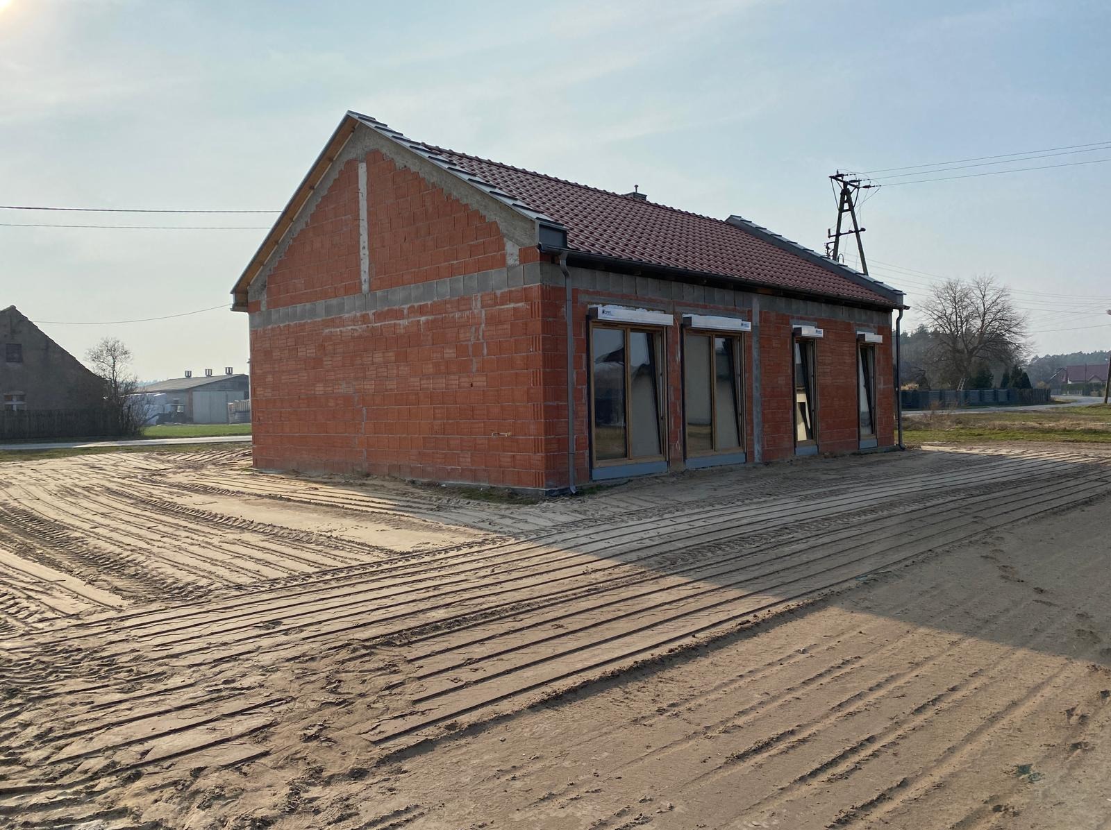 Surowy, ceglany budynek w trakcie budowy z oknami bez szyb, na piaszczystym terenie z wyraźnymi śladami opon. Widoczne elementy konstrukcyjne i dach z czerwonej dachówki.