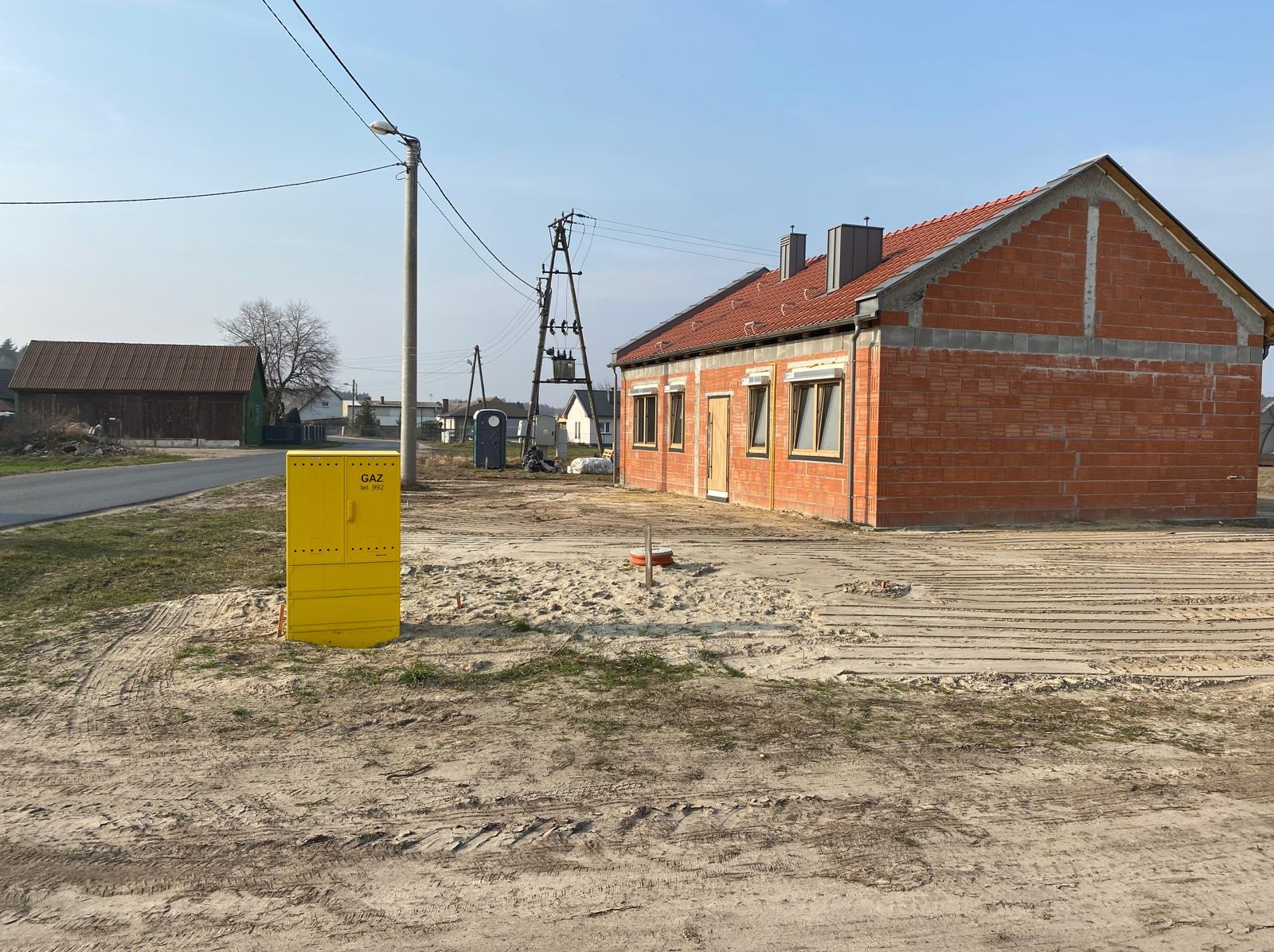 Budowa domu z czerwonej cegły w trakcie realizacji, widoczna instalacja gazowa, teren budowy z piaskiem i śladami opon. Dom z dachem z czerwonej dachówki.