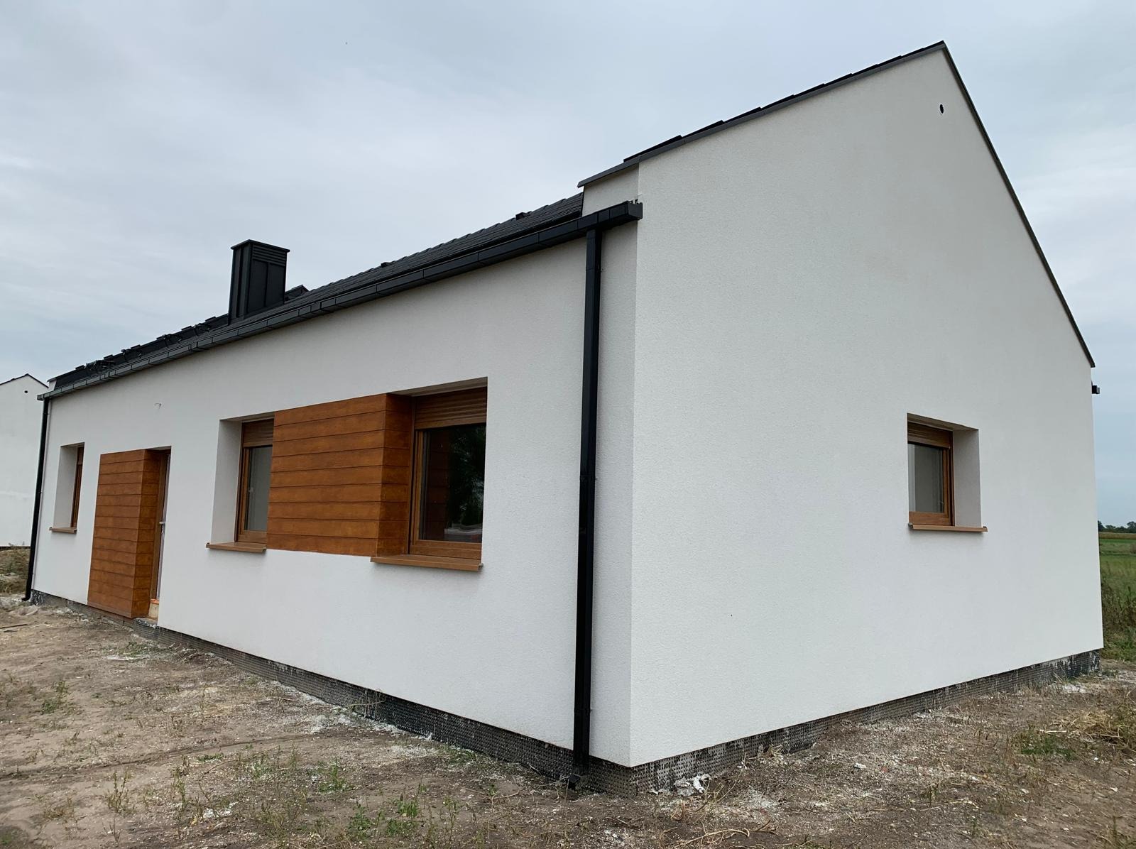 Nowoczesny, parterowy dom z białym tynkiem i drewnianymi elementami okiennymi. Widoczny fragment dachu z czarną dachówką i rynnami. Dom w stanie surowym zamkniętym.