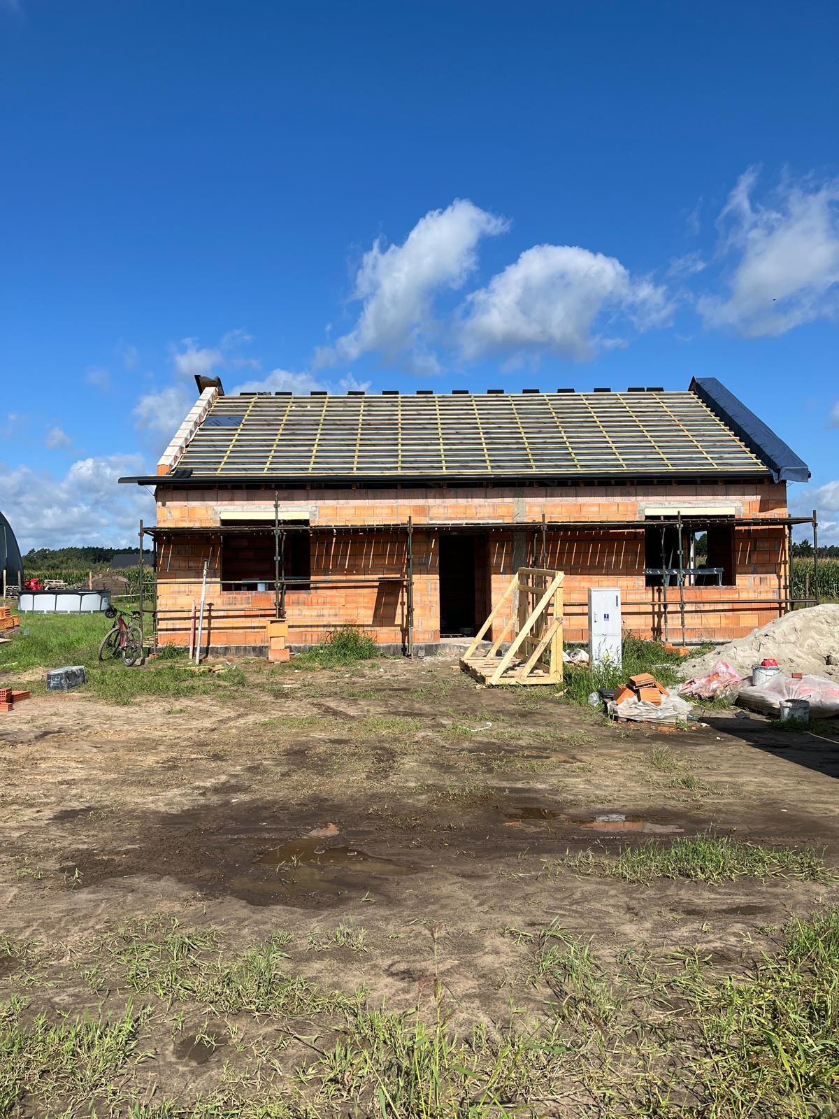 Surowy, ceglany dom w trakcie budowy, z widocznym szkieletem dachu i rusztowaniami. Na pierwszym planie teren budowy z elementami materiałów budowlanych.