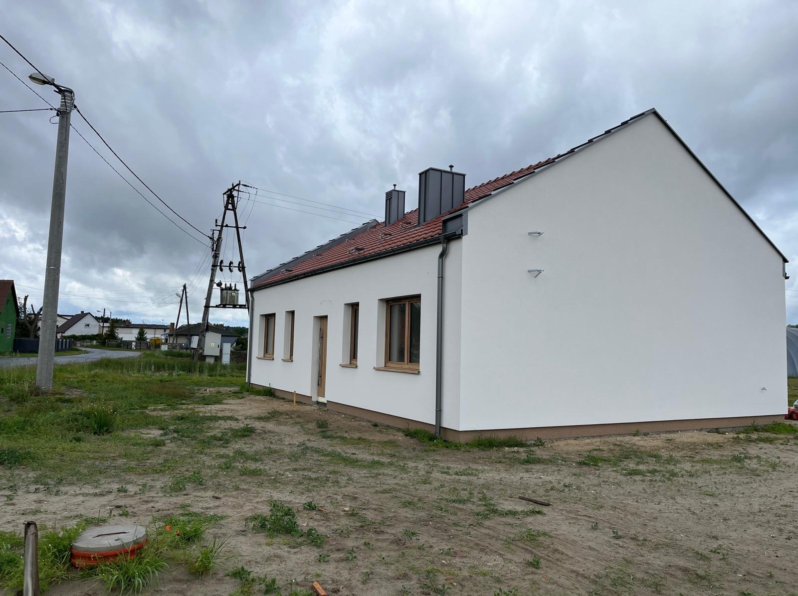 Nowo wybudowany, parterowy dom z białymi ścianami i brązową dachówką, widoczny z boku na tle pochmurnego nieba i słupa energetycznego.