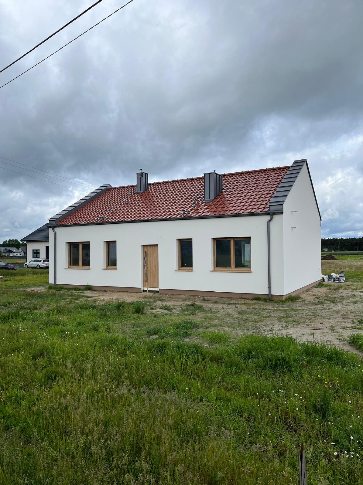 Nowoczesny, parterowy dom z czerwoną dachówką i szarymi kominami, otoczony zielonym trawnikiem pod pochmurnym niebem. Widoczne detale wykończenia.