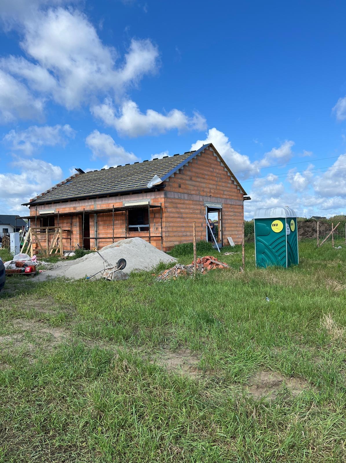 Budowa domu z czerwonej cegły w trakcie realizacji, widoczne rusztowania, sterta piasku i przenośna toaleta na trawiastym terenie w słoneczny dzień.