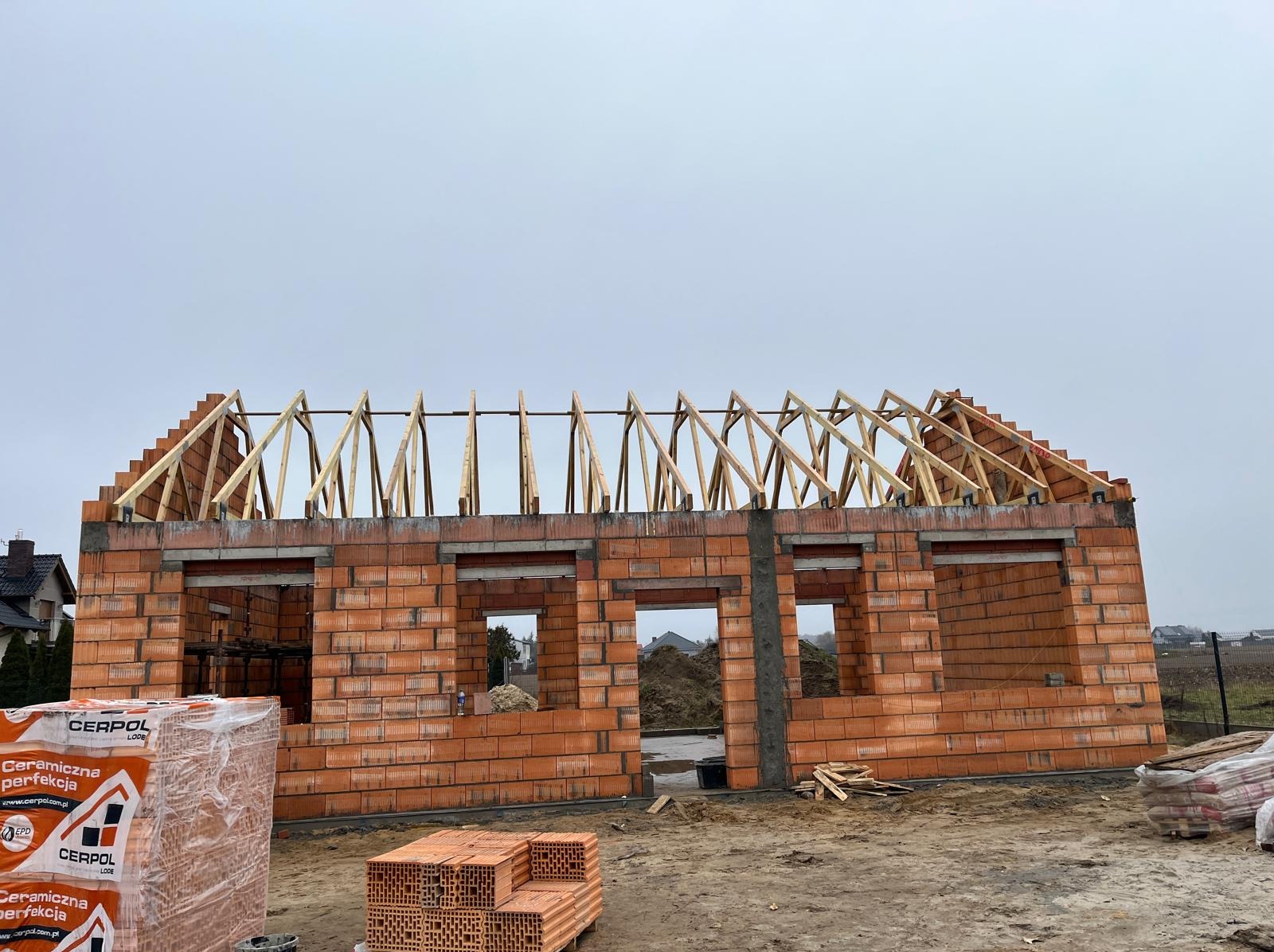 Wznoszenie domu z czerwonej cegły ceramicznej, widoczne ściany z otworami okiennymi i drewniana konstrukcja dachu w trakcie budowy. Na pierwszym planie paleta z cegłami.