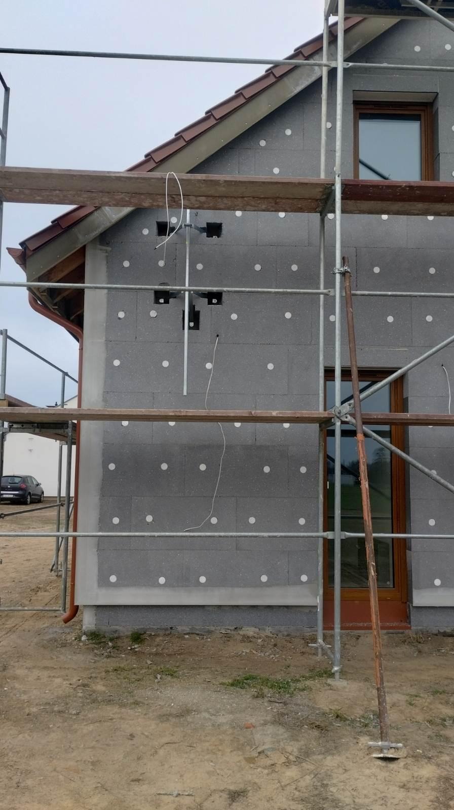 Budowa domu: ściana z szarego bloczka ocieplona styropianem, widoczne rusztowanie i wystające przewody. Etap prac wykończeniowych, widok z zewnątrz.