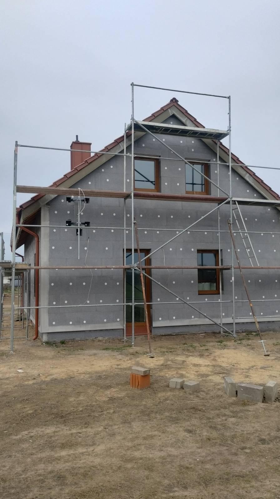 Dom w trakcie ocieplania, widoczne rusztowania, szara elewacja z białymi kołkami. Częściowo ocieplony budynek z brązowymi oknami i dachem.