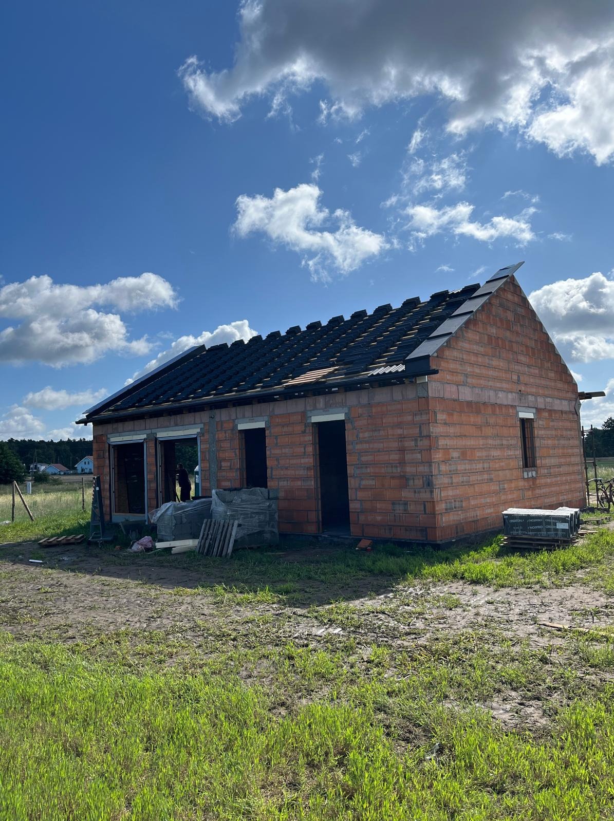 Niedokończony dom z czerwonej cegły z częściowo ułożonym dachem z czarnej dachówki, na tle błękitnego nieba z chmurami i zielonej trawy.