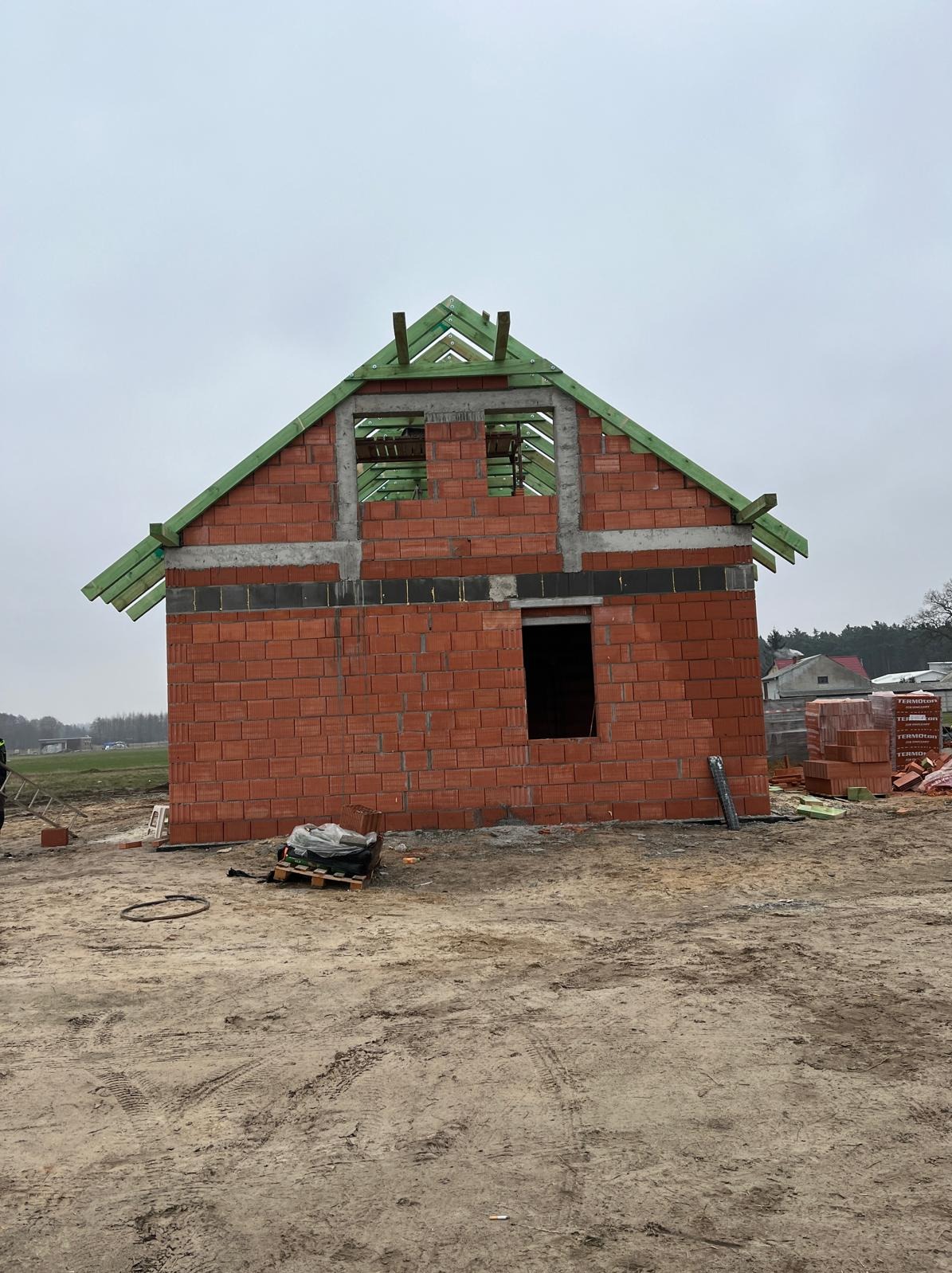 Surowy stan domu z czerwonej cegły z zieloną konstrukcją dachu w trakcie budowy. Widoczne palety z materiałami budowlanymi i ziemiste podłoże.