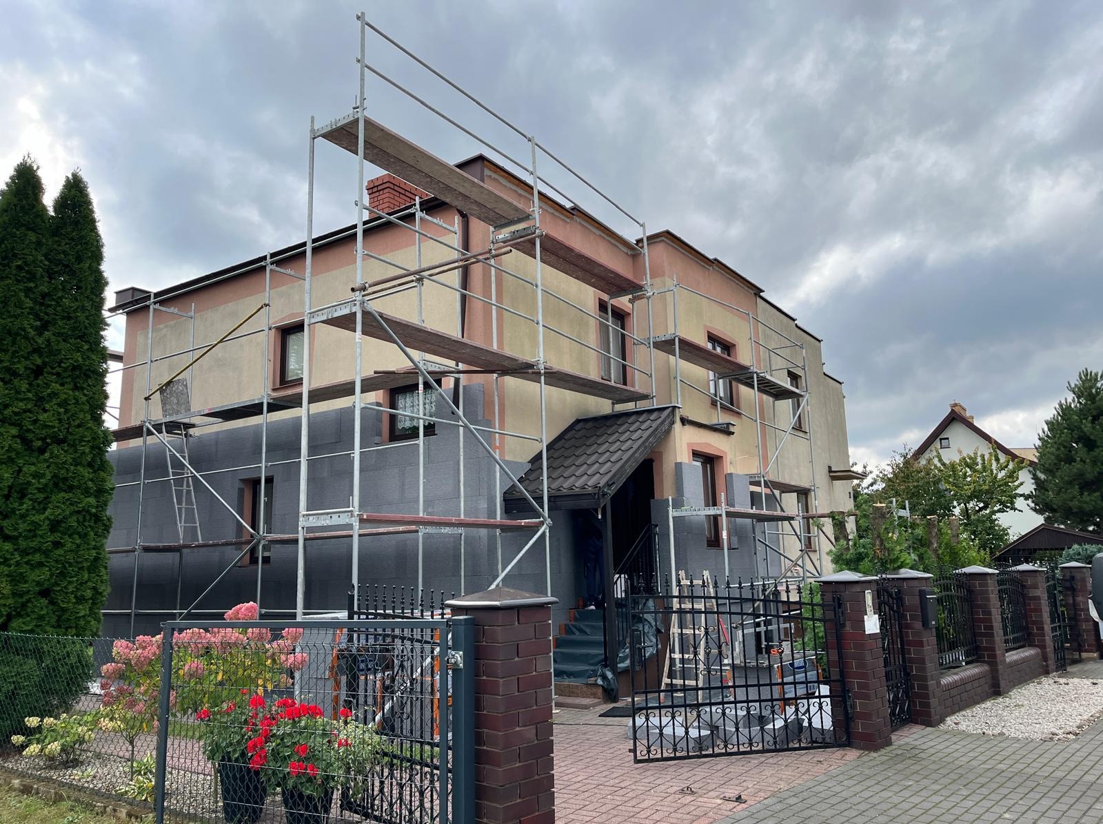 Ocieplanie elewacji domu jednorodzinnego z użyciem rusztowania. Widoczne płyty izolacyjne na ścianach. Częściowo ocieplony budynek z ciemnymi oknami i ogrodzeniem.
