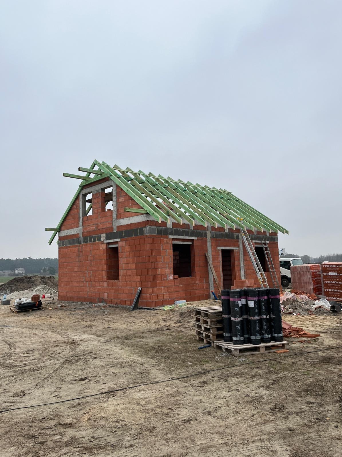 Surowy stan domu z czerwonej cegły z zieloną konstrukcją dachu, drabina oparta o ścianę, materiały budowlane na ziemi, pochmurne niebo w tle.
