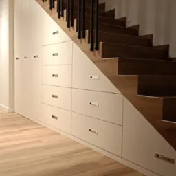 Białe szafki i szuflady w zabudowie pod drewnianymi schodami z czarną balustradą, jasna drewniana podłoga.