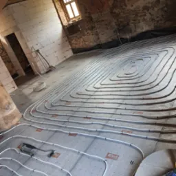 Montaż ogrzewania podłogowego w pomieszczeniu z surowymi ścianami z cegły i pustaków, widoczne rury grzewcze ułożone w pętle na izolacji z nadrukowanym logo, narzędzia montażowe obok.