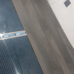 Montaż folii grzewczej pod panele podłogowe o ciemnym odcieniu drewna, widoczne przewody elektryczne i gniazdko w ścianie.