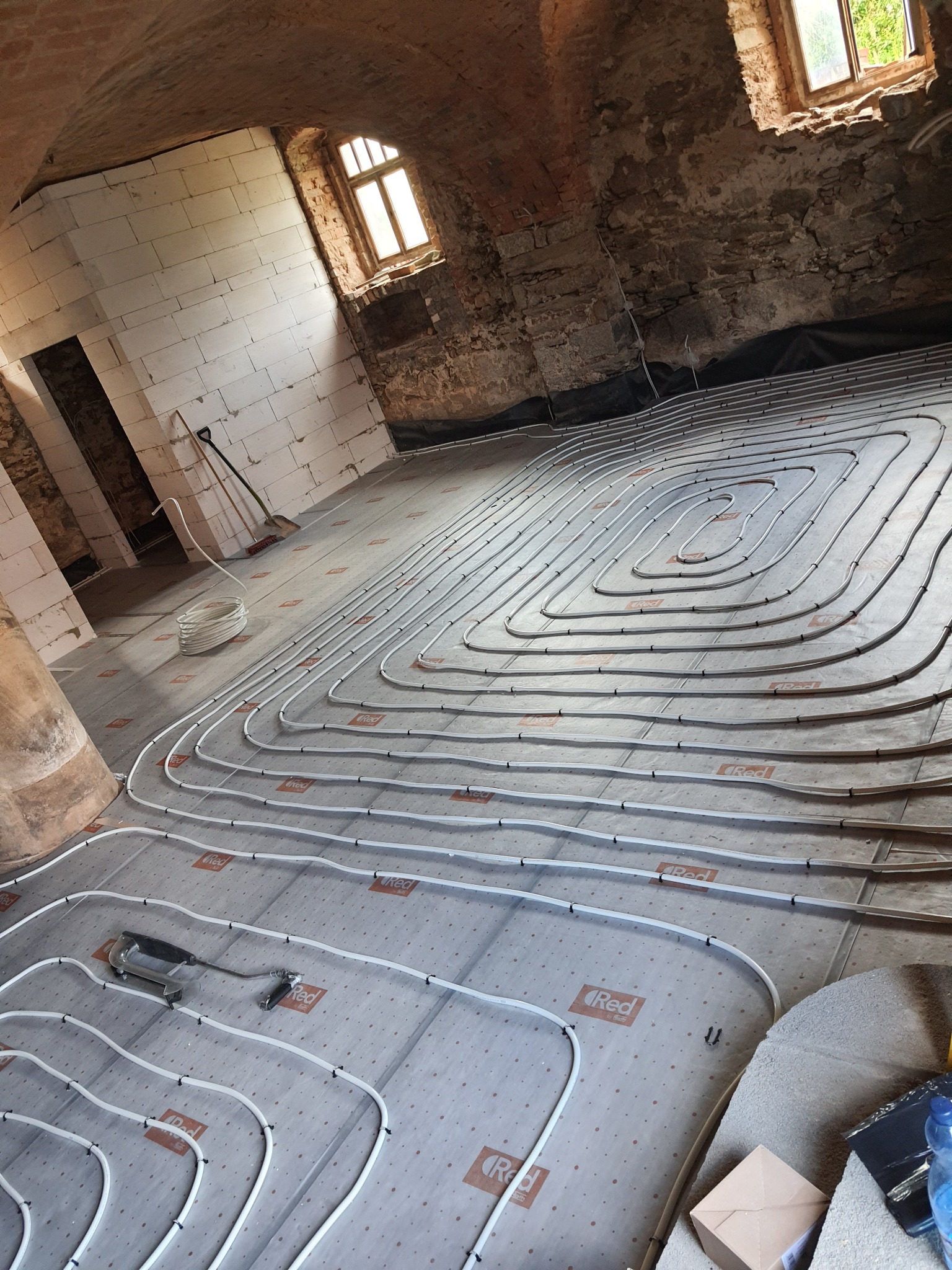 Montaż ogrzewania podłogowego w pomieszczeniu z surowymi ścianami z cegły i pustaków, widoczne rury grzewcze ułożone w pętle na izolacji z nadrukowanym logo, narzędzia montażowe obok.