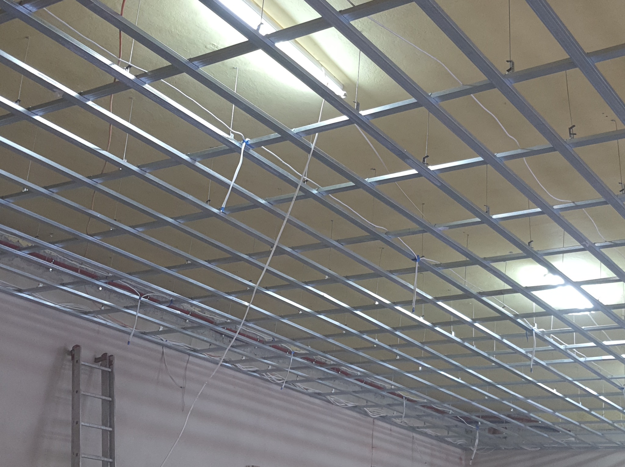 Metalowa konstrukcja sufitowa w trakcie montażu, widoczne kable i rury instalacyjne, drabina oparta o ścianę w tle.