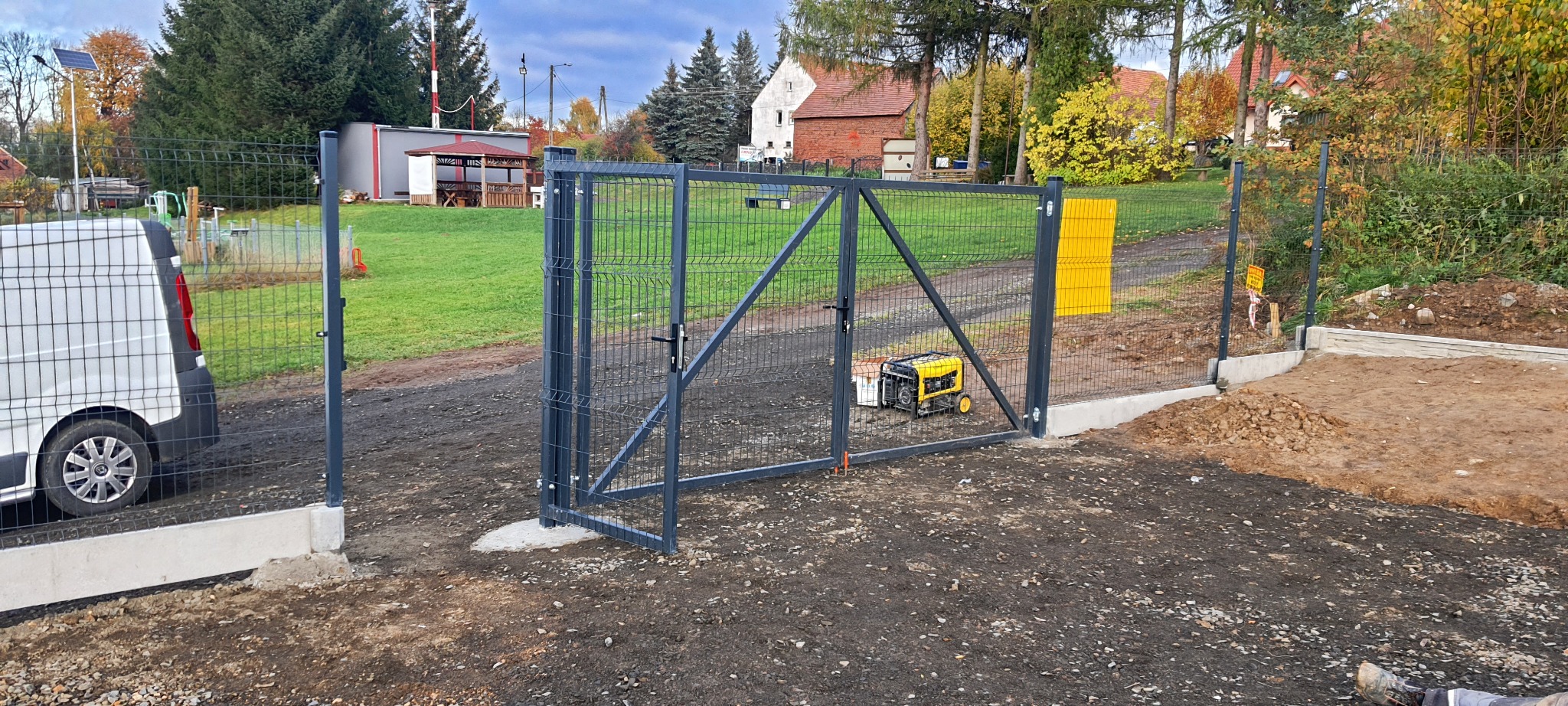 Nowo zamontowana, stalowa brama panelowa z podmurówką w Gryfowie Śląskim. Widoczny agregat prądotwórczy i częściowo biały samochód dostawczy. W tle budynki mieszkalne i zieleń.