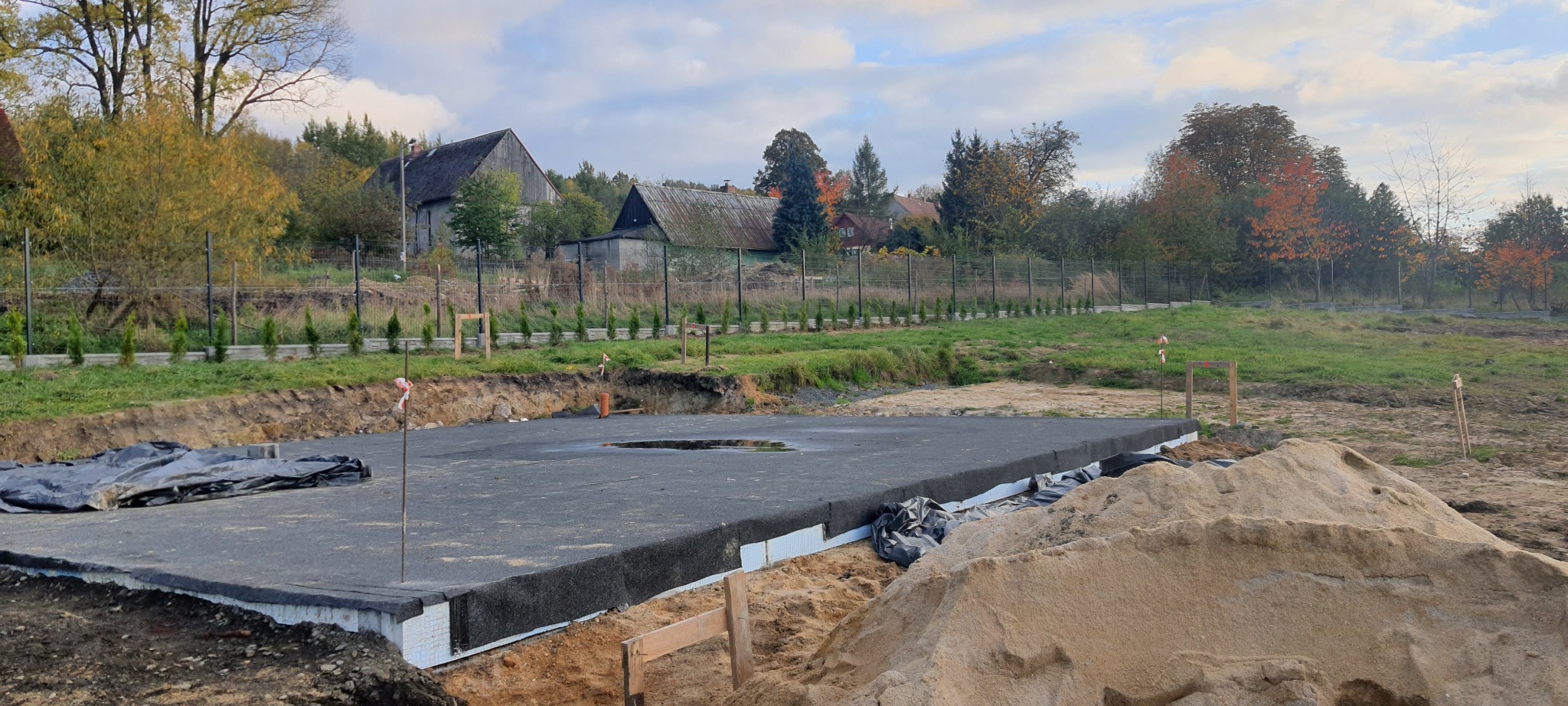 Przygotowany fundament pod budynek, zaizolowany styropianem i folią, z widocznym wykopem i stertą piasku w tle krajobraz wiejski z drzewami i zabudowaniami.