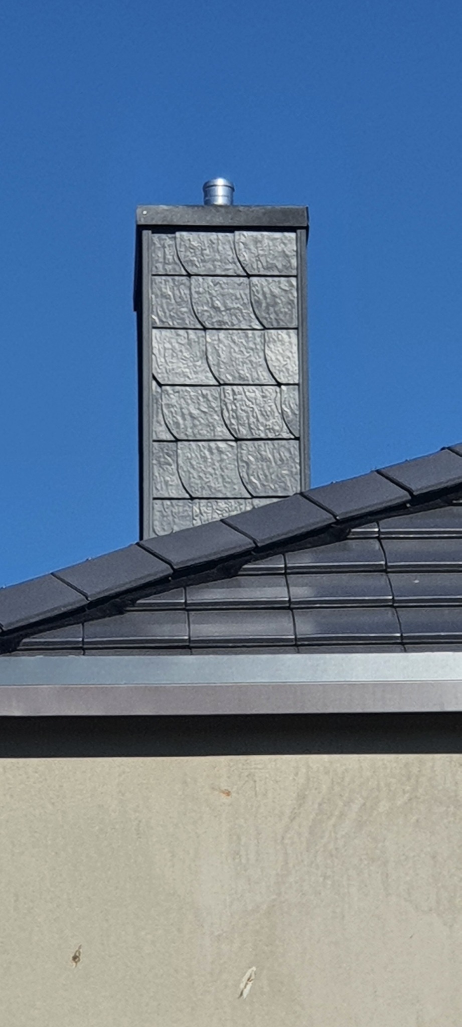 Kominek z szarej łupki na tle błękitnego nieba, widok z dołu, fragment dachu z ciemnej dachówki.