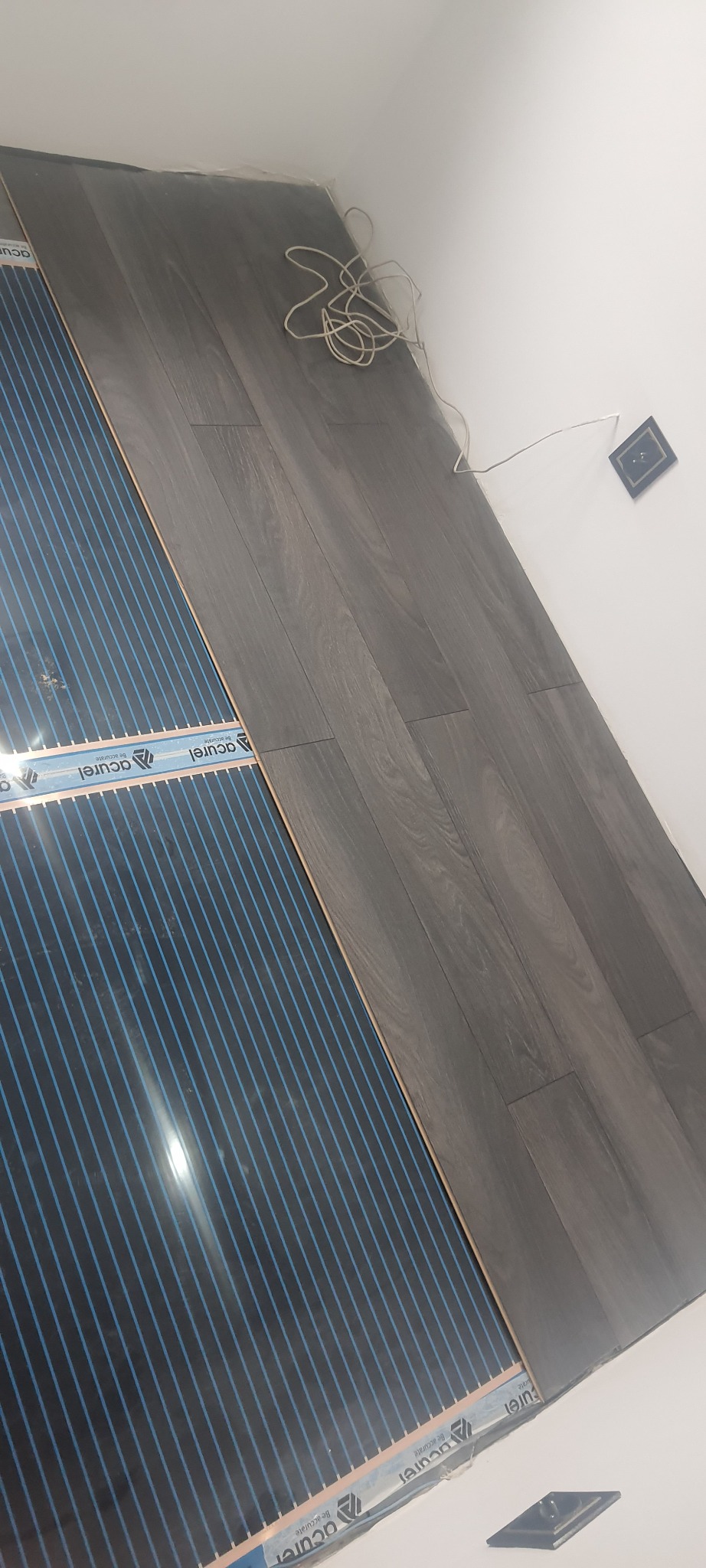 Montaż folii grzewczej pod panele podłogowe o ciemnym odcieniu drewna, widoczne przewody elektryczne i gniazdko w ścianie.