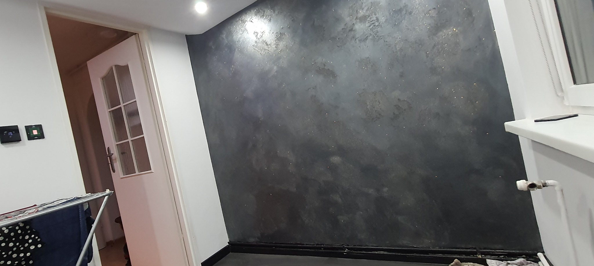 Ściana w pokoju wykończona dekoracyjnym tynkiem w odcieniach szarości z delikatnymi złotymi akcentami, widoczna biała stolarka drzwiowa i fragment suszarki na pranie.