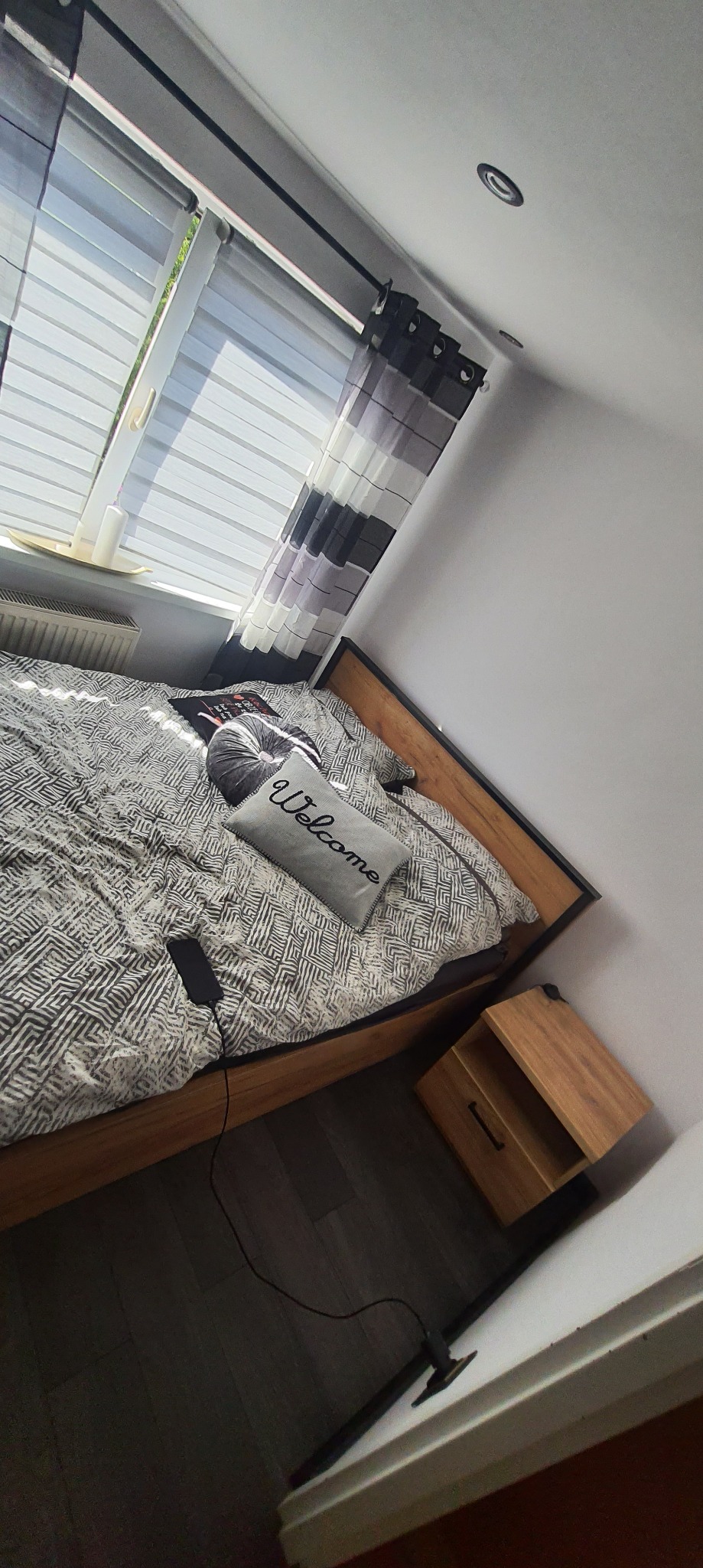 Sypialnia z drewnianym łóżkiem, wzorzystą pościelą, poduszką z napisem 'Welcome' i szafką nocną z drewna, oświetlona przez okno z żaluzjami.